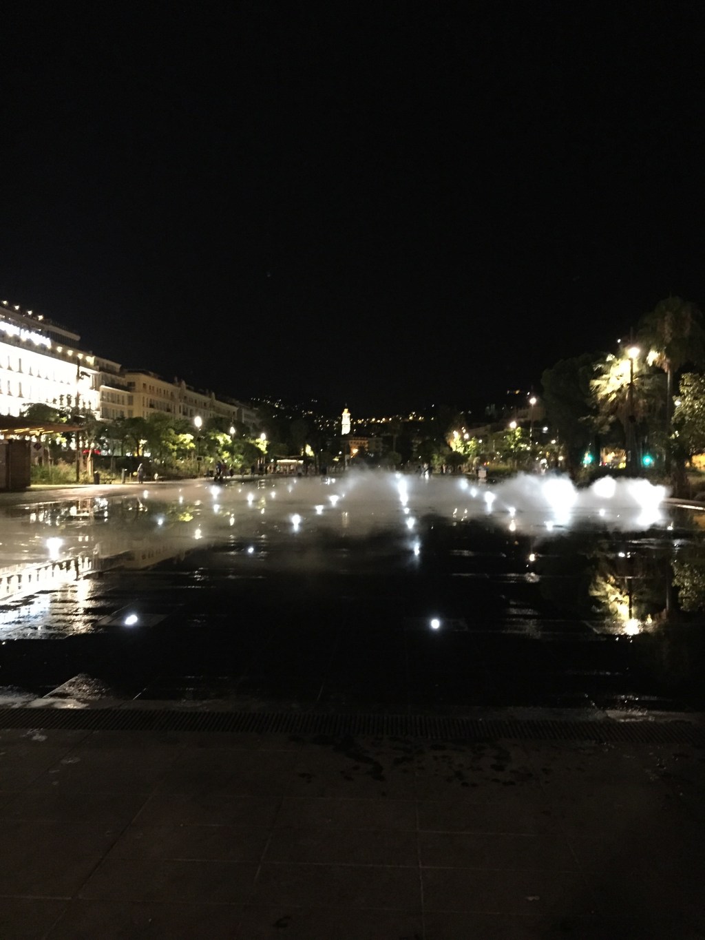 The mirror fountain on the Promenade du Paillon at night, low jets throwing mist across the wet stone with the lit facades behind.