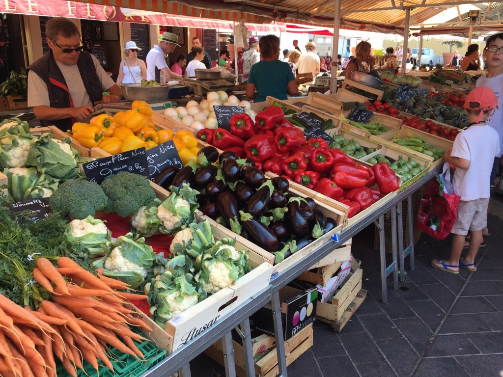 A produce stall at the Cours Saleya market piled with yellow and red peppers, aubergines, carrots and cauliflower under a striped awning.