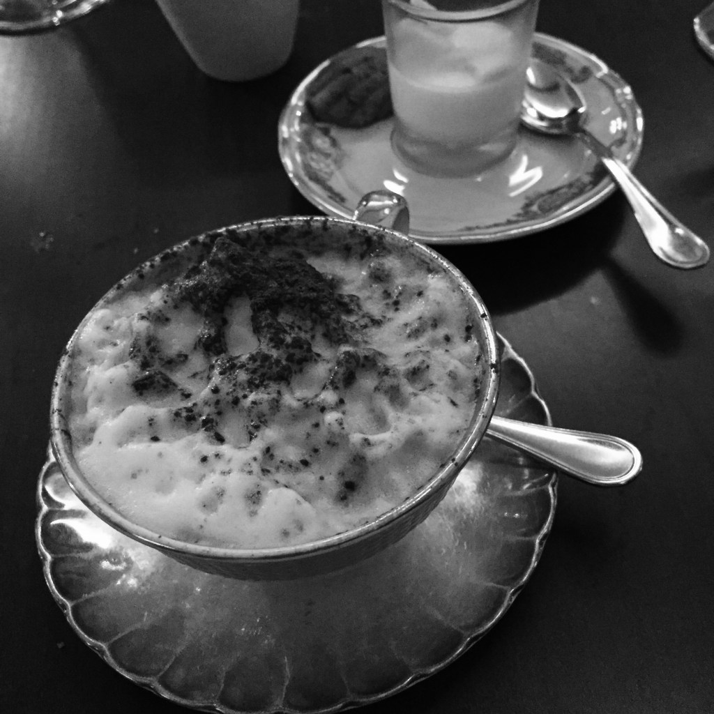 A cappuccino dusted with chocolate in a scalloped porcelain cup, a lemon dessert in a glass on the saucer behind it.