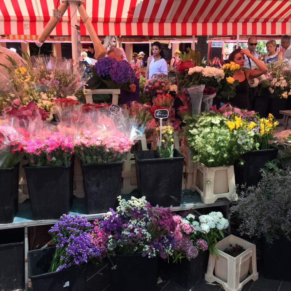 A flower stall under a red and white striped awning, buckets of pink, yellow and purple blooms crowding the pavement.