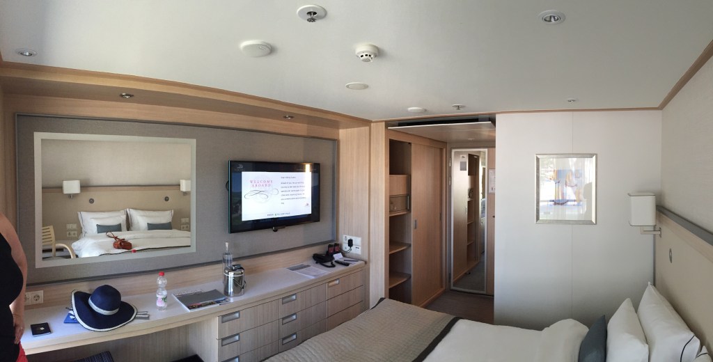 A river cruise cabin: bed, mirrored wall, television and desk, with a straw hat set down on the sideboard.