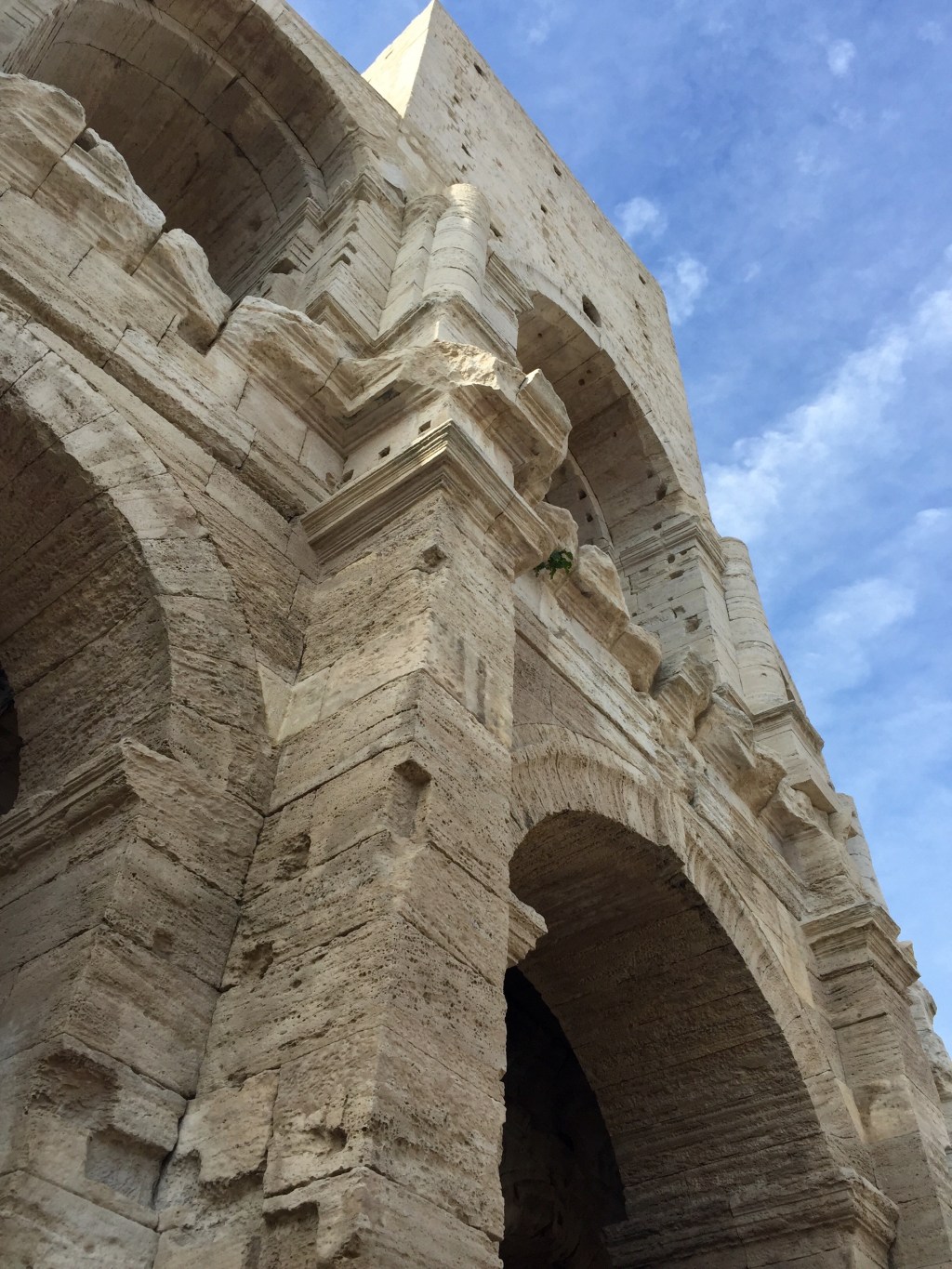 The Roman amphitheatre at Arles seen from below, two tiers of pale stone arches against a blue sky.