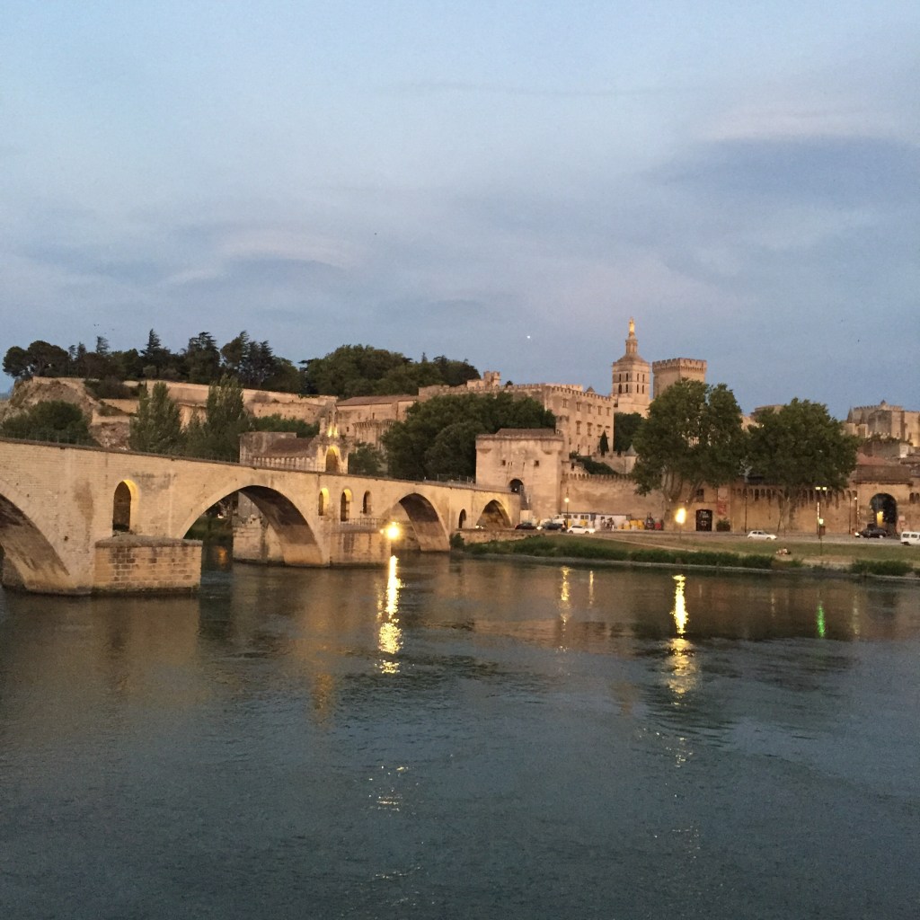 The Pont Saint-Bénézet and the Palais des Papes at dusk, seen across the Rhône with lights reflecting on the water.
