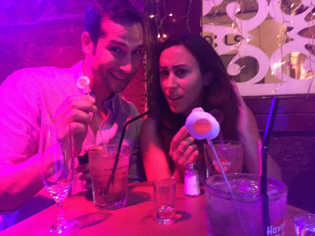 Two people at a bar table under pink and purple light, tall cocktails in front of them.