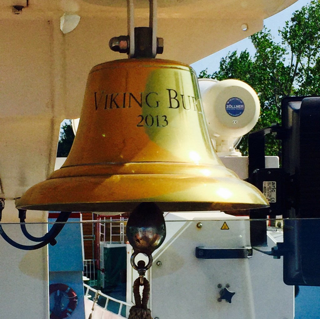 The ship&rsquo;s brass bell, engraved VIKING BURI 2013, mounted under the deckhead with the riverbank beyond.