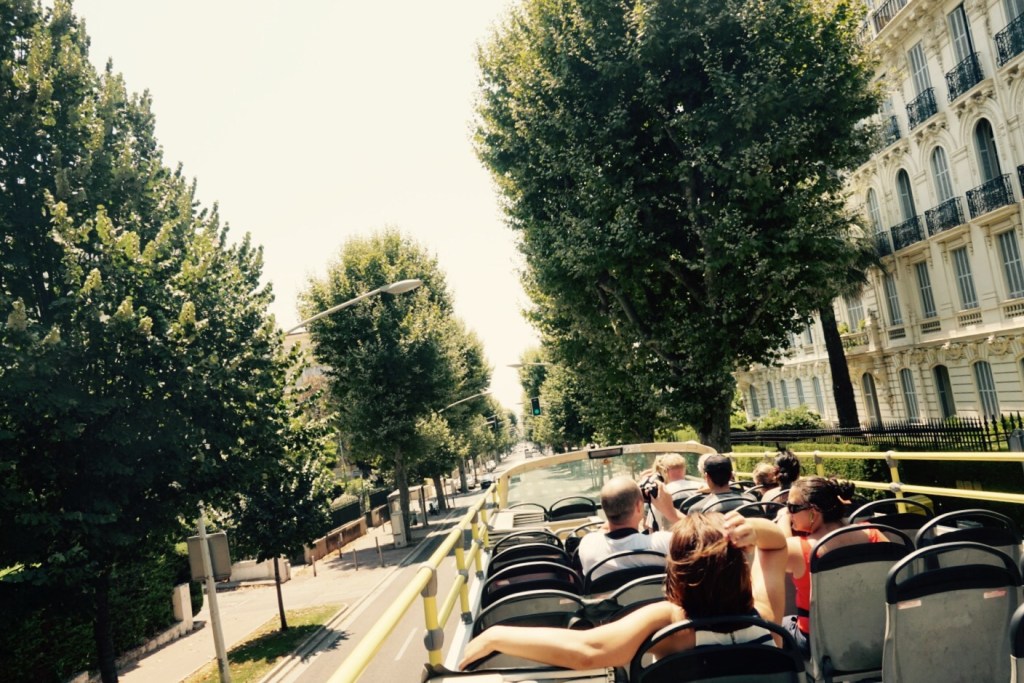 The open top deck of a sightseeing bus running down a broad avenue in Nice between plane trees and pale apartment blocks.