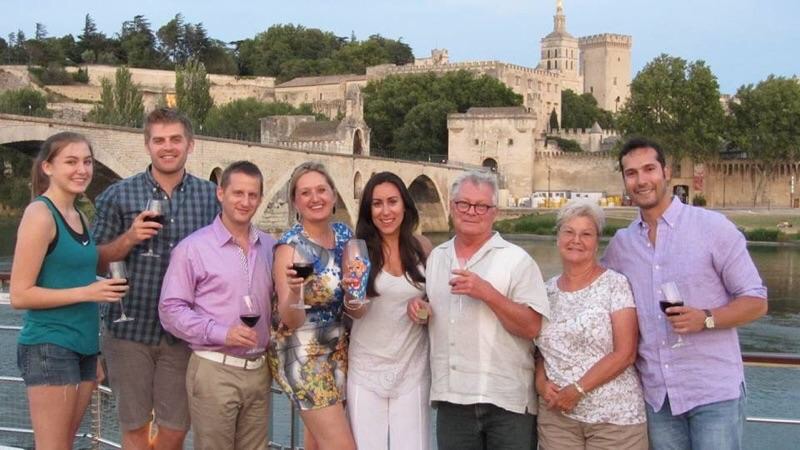 Eight passengers lined up with glasses of wine on the ship&rsquo;s top deck, the Palais des Papes and the broken Pont Saint-Bénézet behind them across the water.