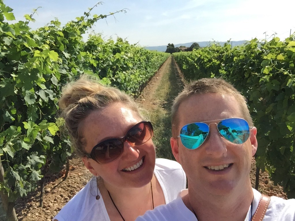 Chris and Jen between two rows of vines in full leaf, the row running away behind them to distant hills.