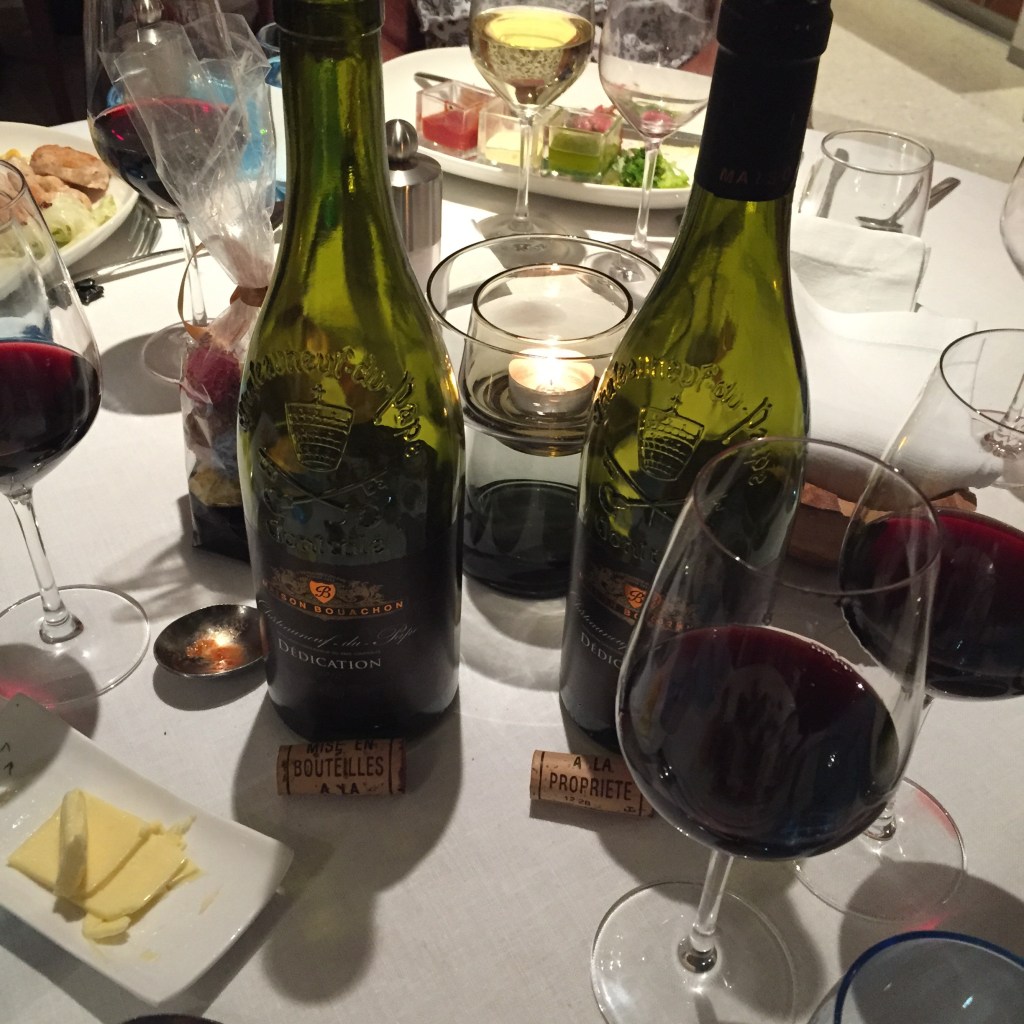 Two bottles of red wine and filled glasses on a white restaurant table, candles and a cheese plate alongside.