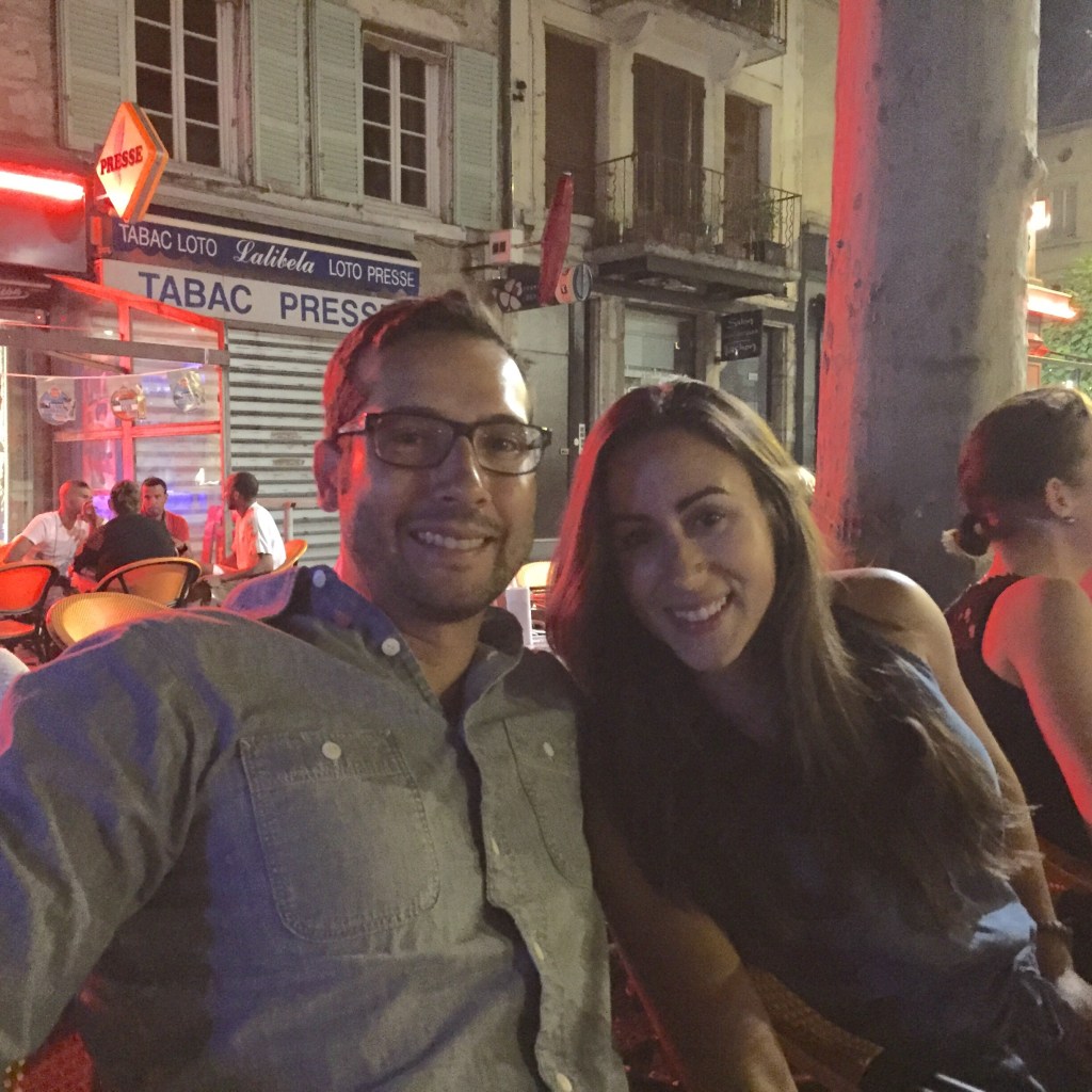 A night street scene, people at outdoor tables under the red neon of a tabac sign.