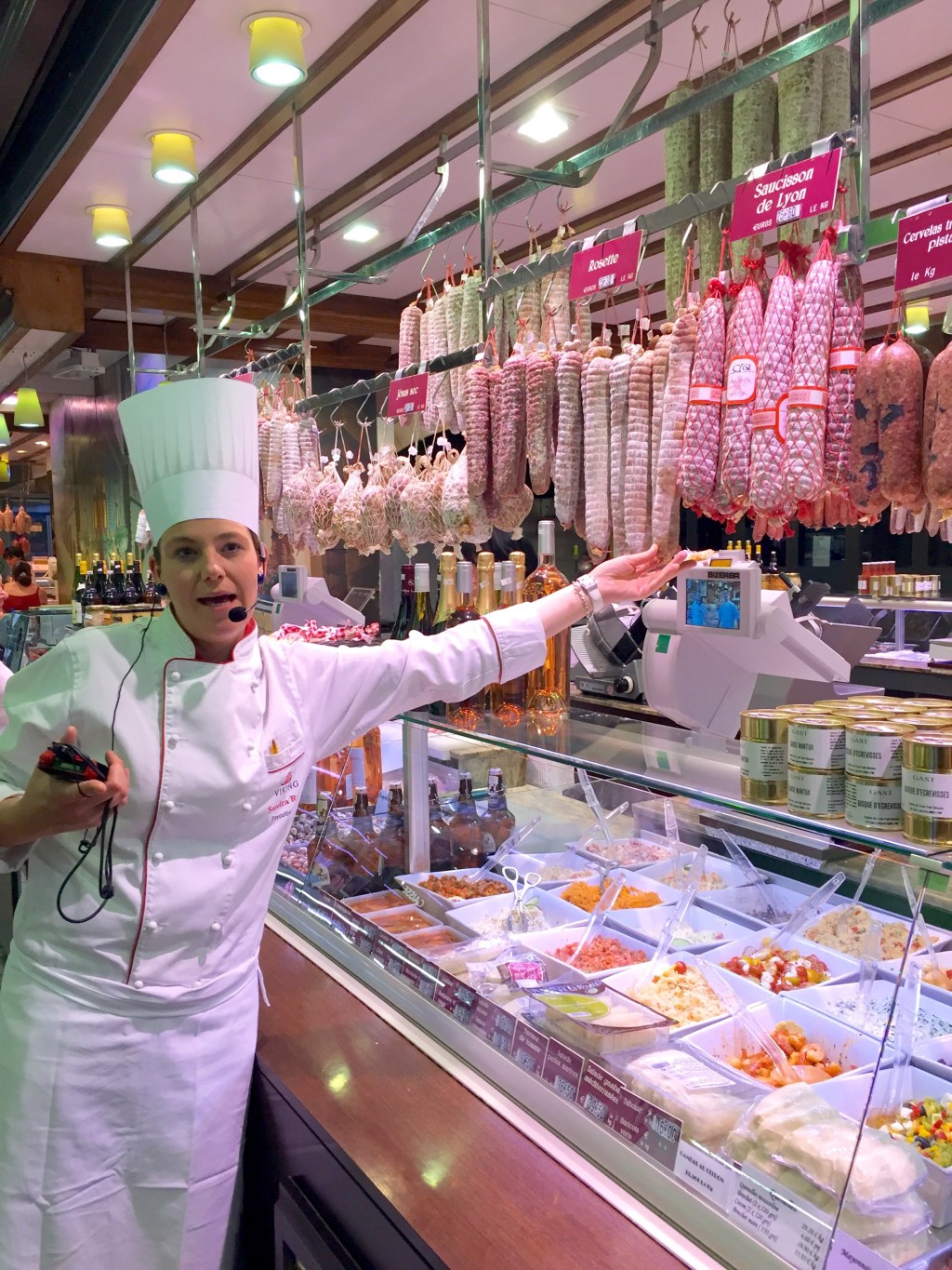 A chef in whites gesturing along a charcuterie counter hung with rows of saucisson, trays of prepared food in the case below.
