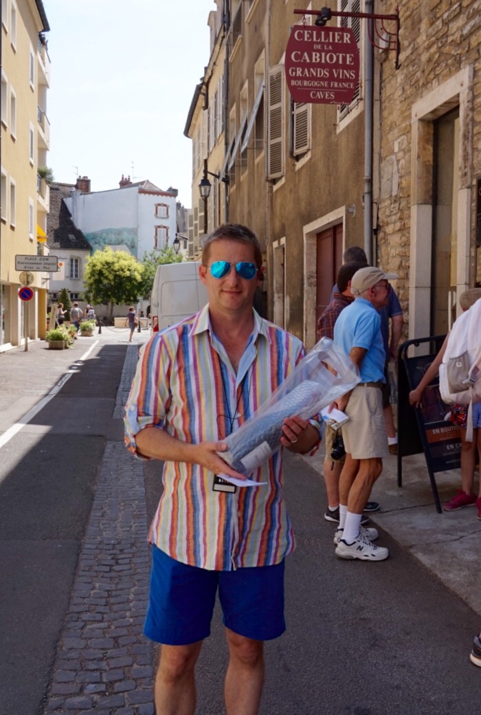 Chris in a striped shirt holding a rolled poster in a Beaune street, a wine cellar sign above the doorway behind him.