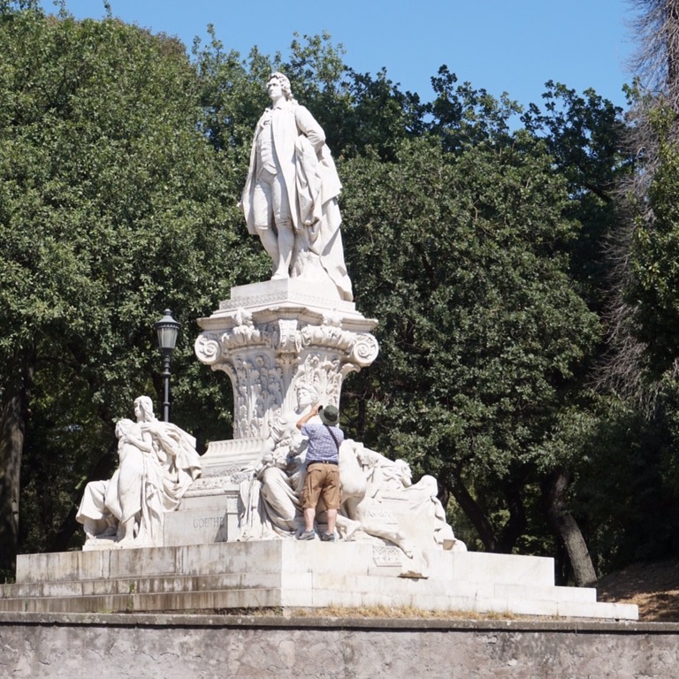 A white marble monument in a Rome park, a man sitting on the steps at its base among the sculpted figures