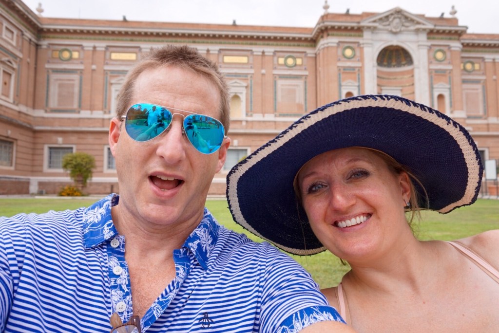 Chris and Jen on the lawn in the Vatican Museums courtyard, Chris in mirrored sunglasses and Jen in a wide navy sun hat