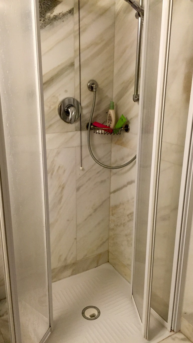 A marble hotel bathroom with a glass-walled shower, a hand shower on a rail and a small red charm hanging beside it