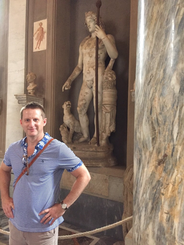 Chris standing beside a marble statue group set in a niche in the Vatican Museums