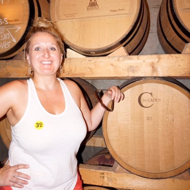 Jen standing beside stacked oak barrels in the bodega cellar, a numbered tour sticker on her top