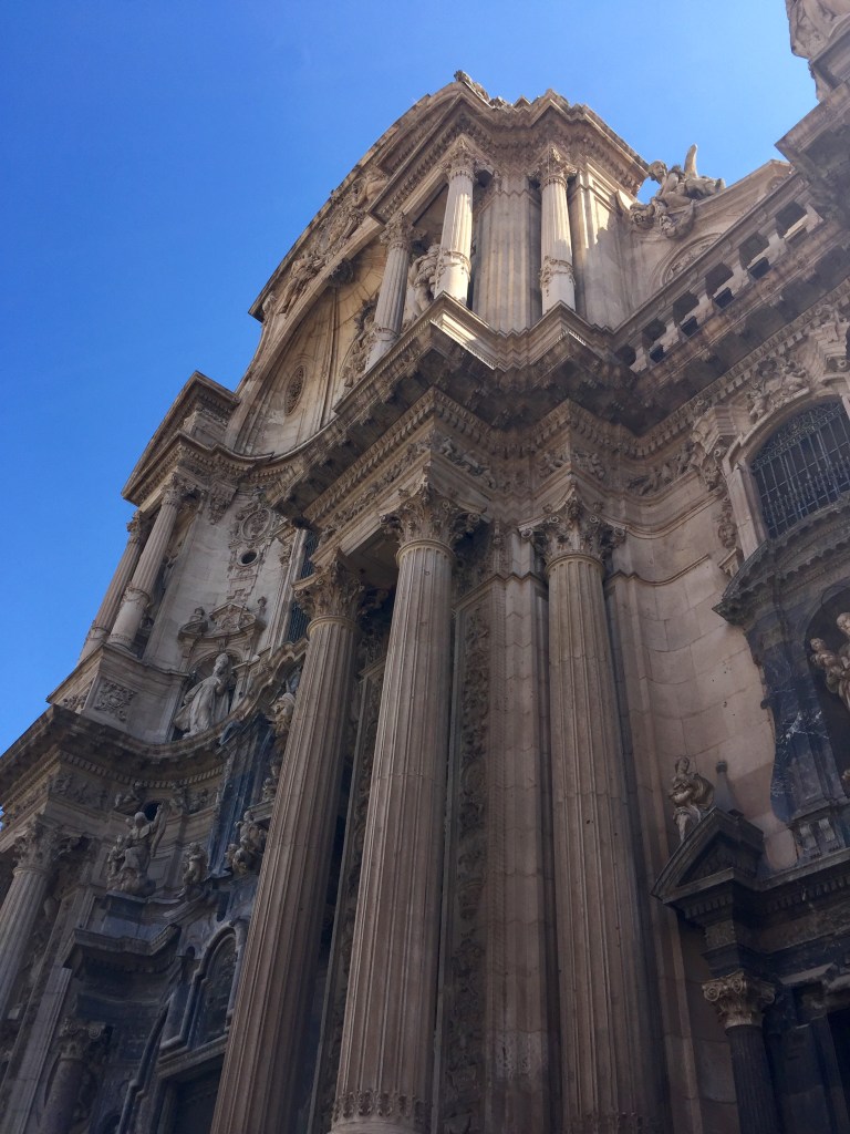 The baroque front of Murcia Cathedral seen from below, columns and statuary against the sky