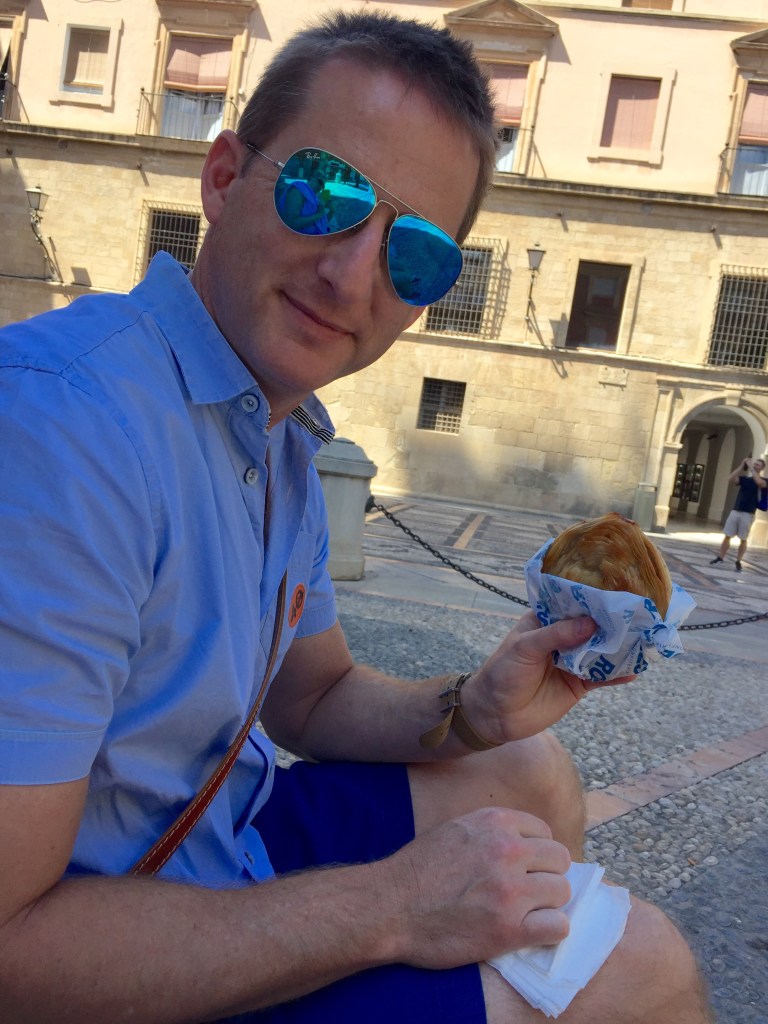 Chris sitting on a step in a Murcia square eating a pastry from its wrapper