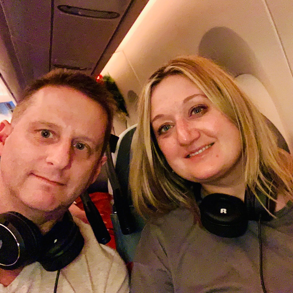 Chris and Jen in headphones, taking a selfie in their seats before the long flight
