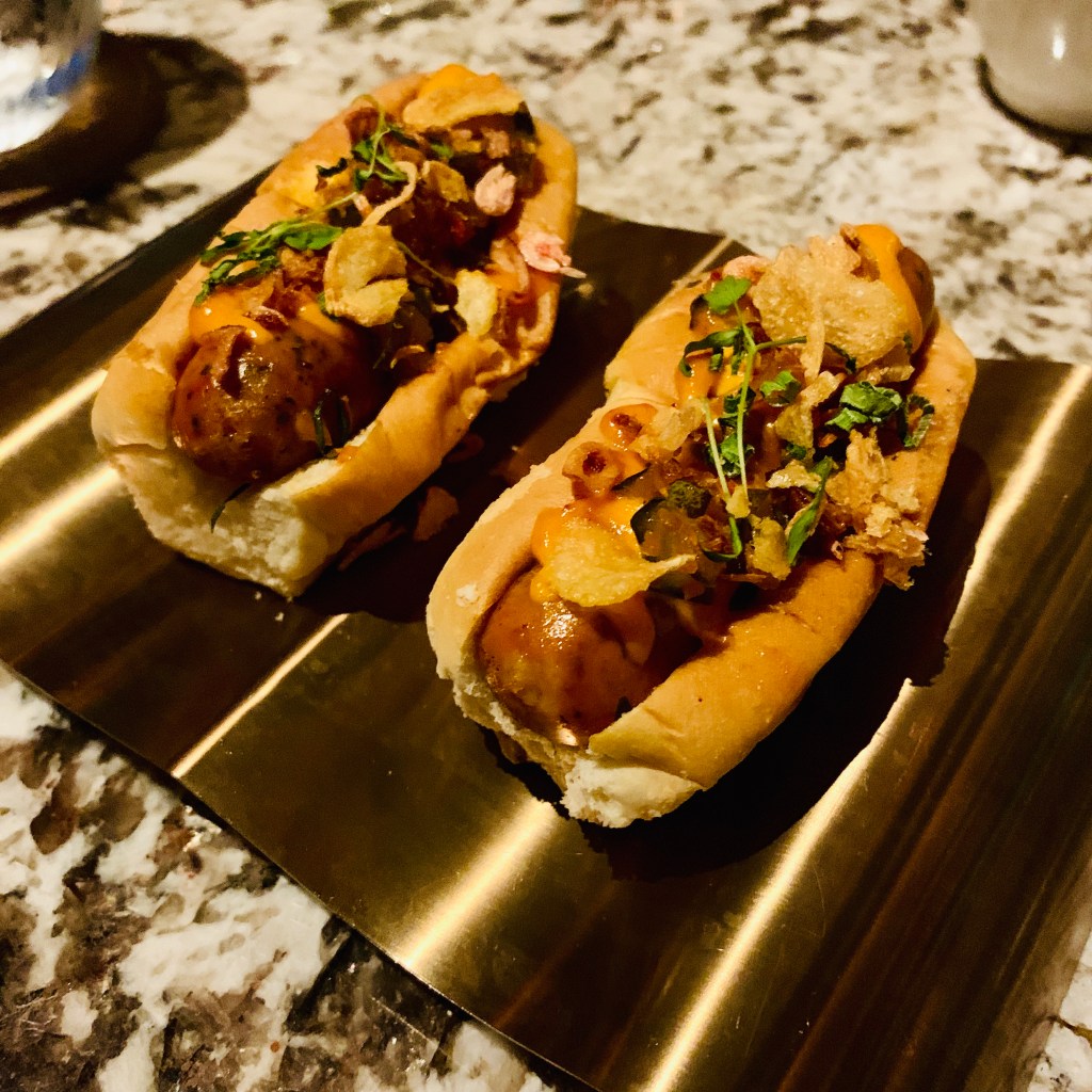 Two loaded hot dogs in gold foil trays, heaped with toppings