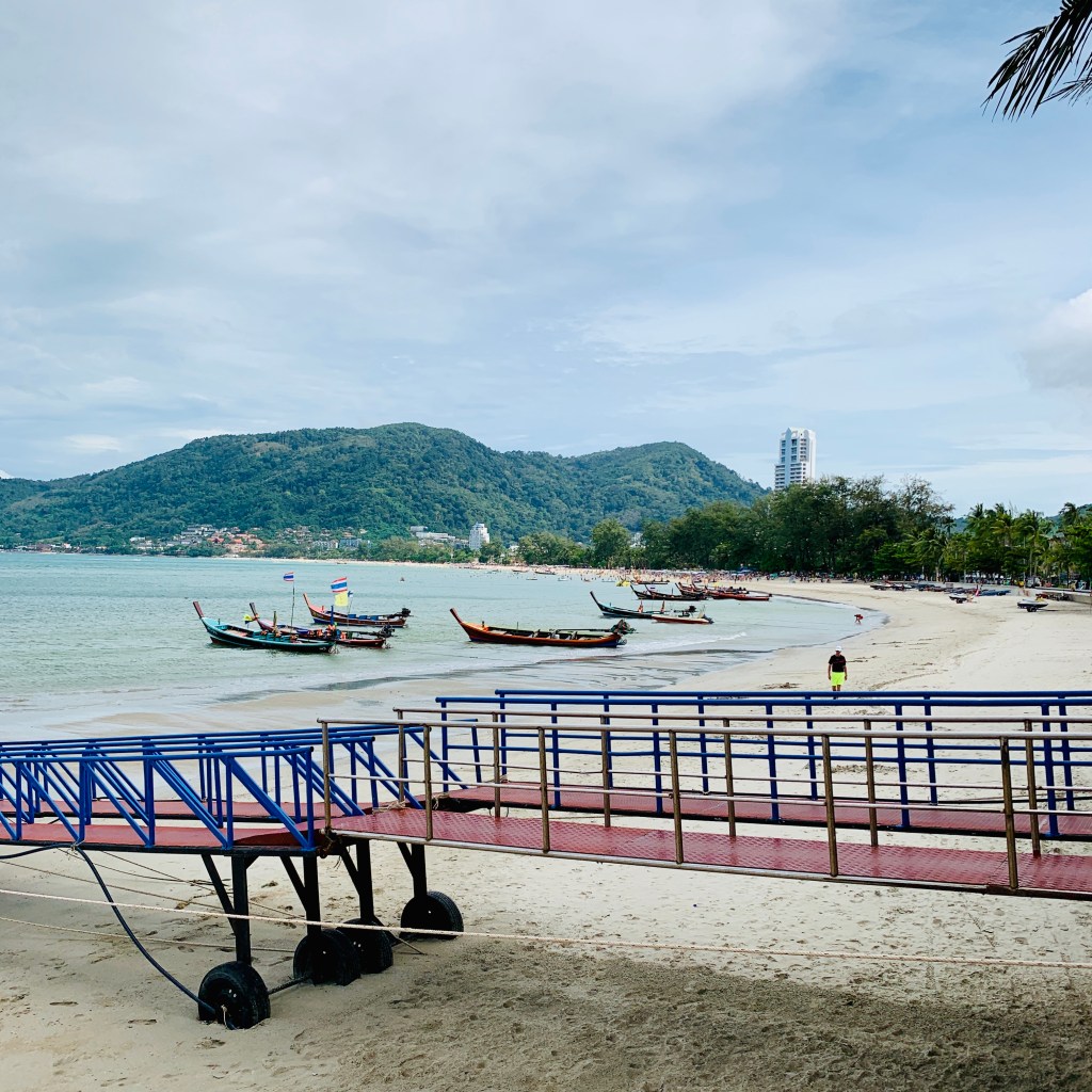 A jetty running out into the bay, longtail boats moored along it under a wooded headland