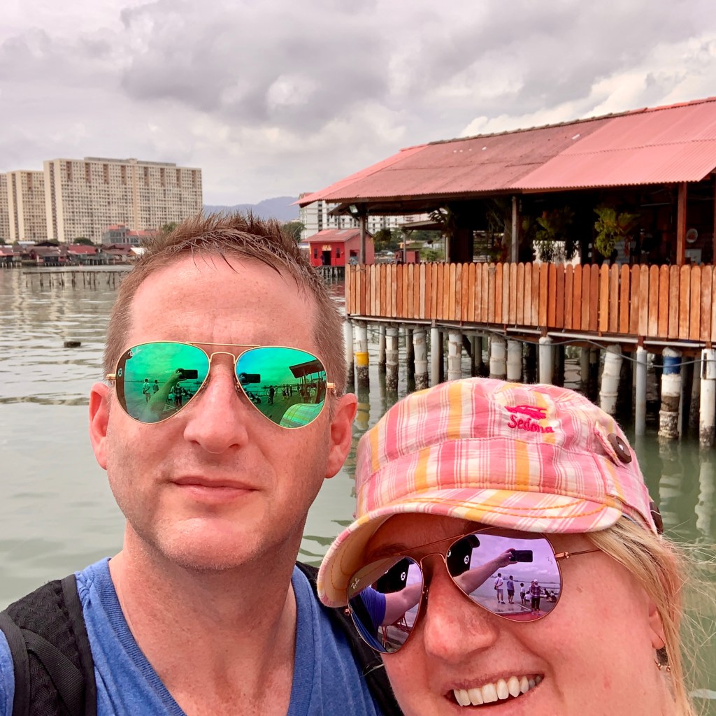 Chris and Jen in sunglasses on the clan jetty, the stilt houses standing over the water behind them