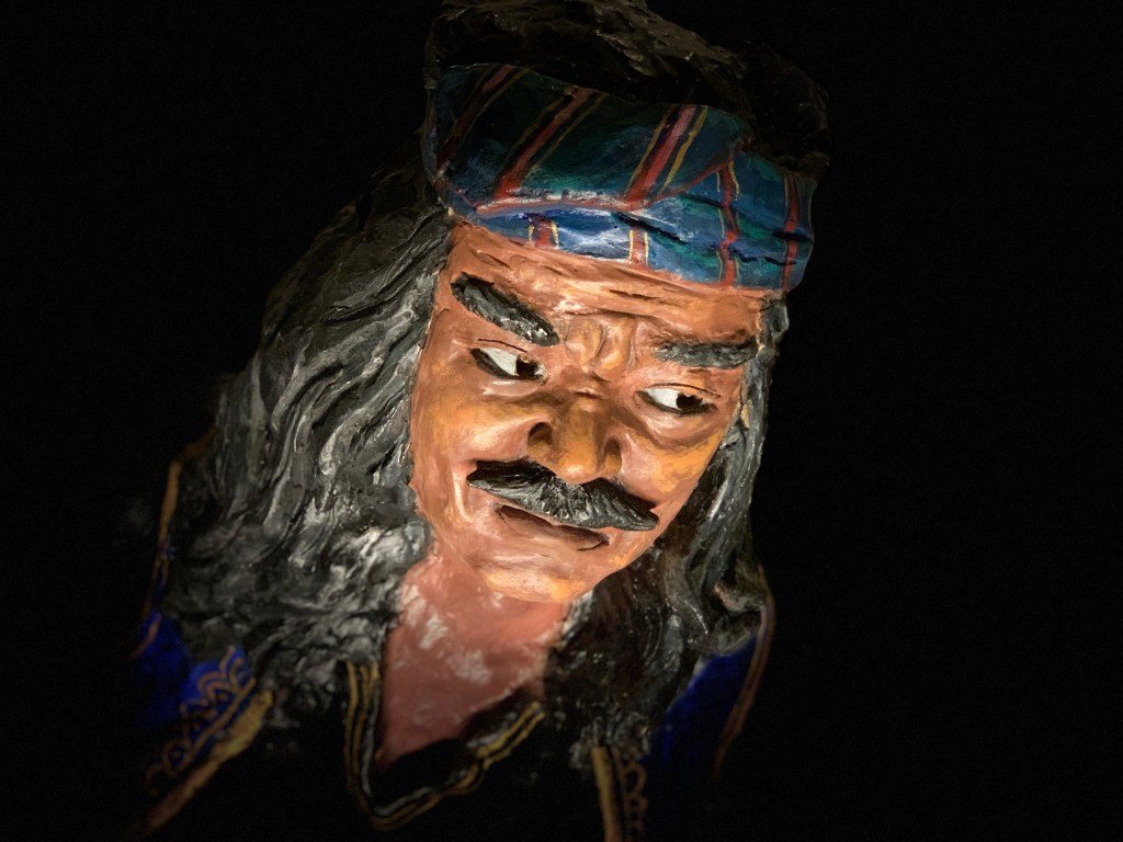 A carved wooden mask with long grey hair, lit against a black museum wall
