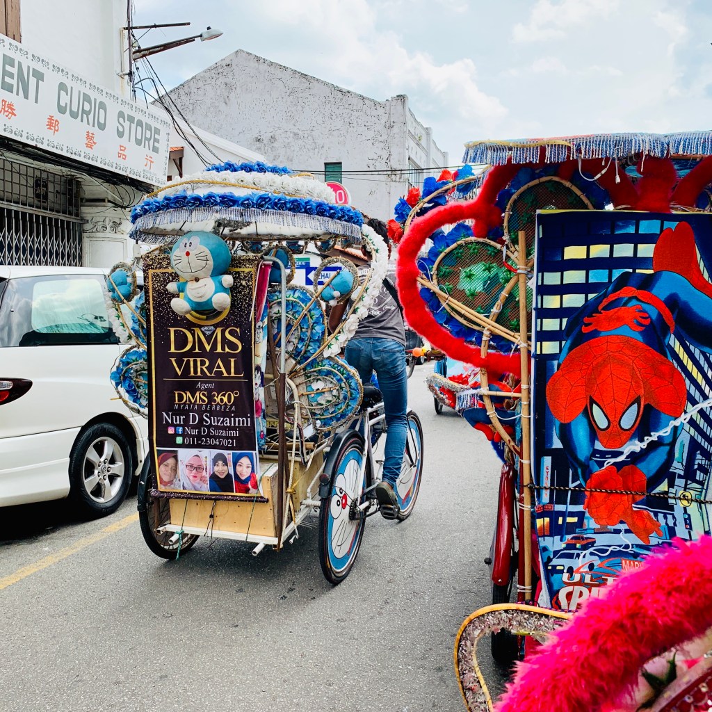 A Malacca trishaw buried under artificial flowers, its driver waiting at the kerb