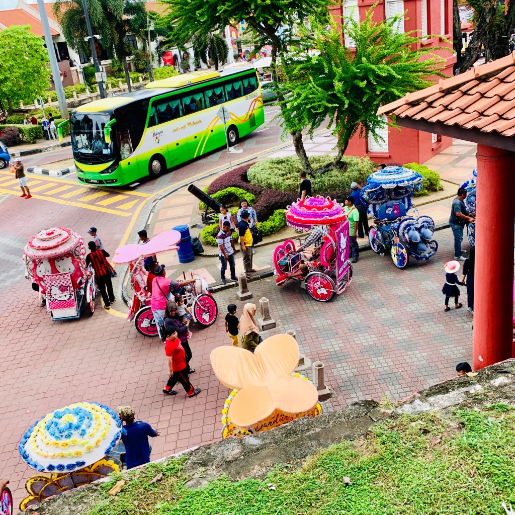 Looking down on a line of decorated trishaws parked along the street, a green bus behind them
