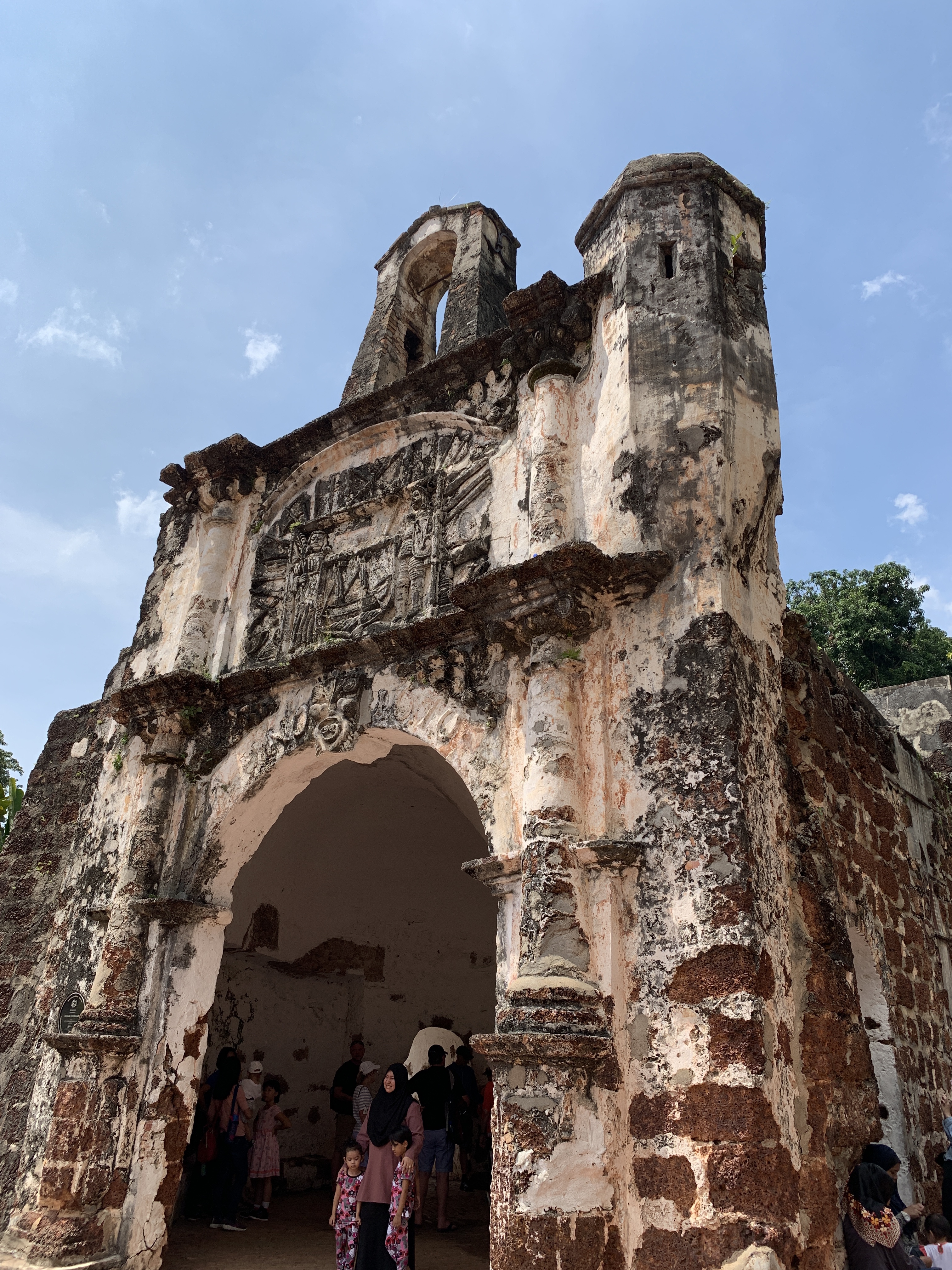 A ruined stone gateway in Malacca, its top ragged with age, visitors standing in the arch