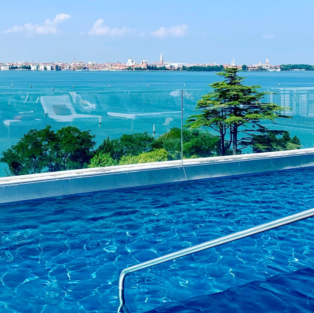 An infinity pool of bright blue water with the Venice skyline low on the horizon across the lagoon beyond the glass balustrade.
