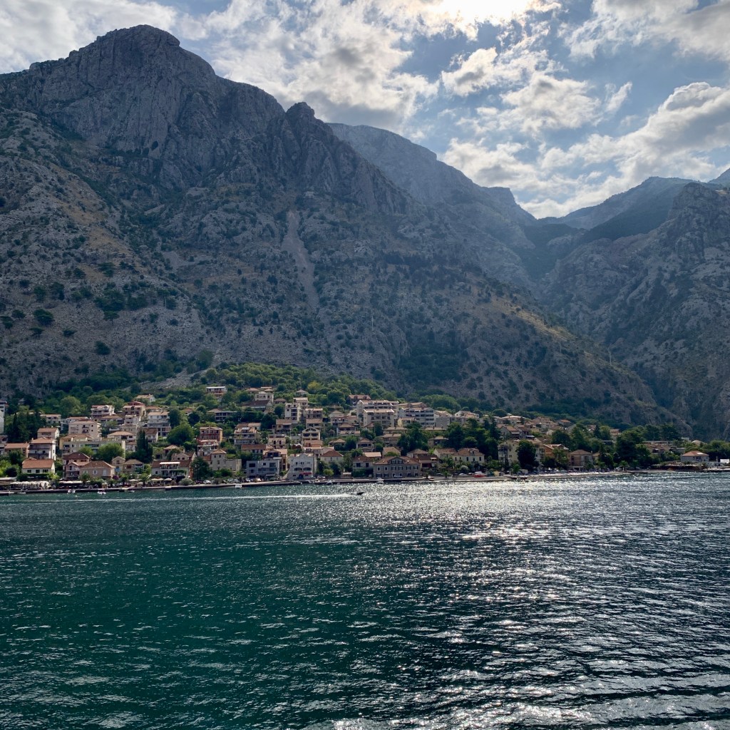 The Bay of Kotor from the water, a line of red-roofed houses strung along the shore beneath a wall of steep grey mountain.
