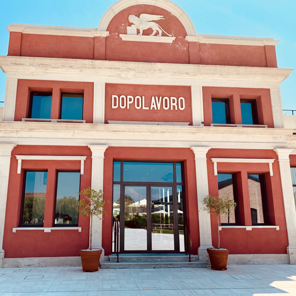 A red and white industrial building with DOPOLAVORO lettering across the facade and a winged lion of Saint Mark in the pediment above.