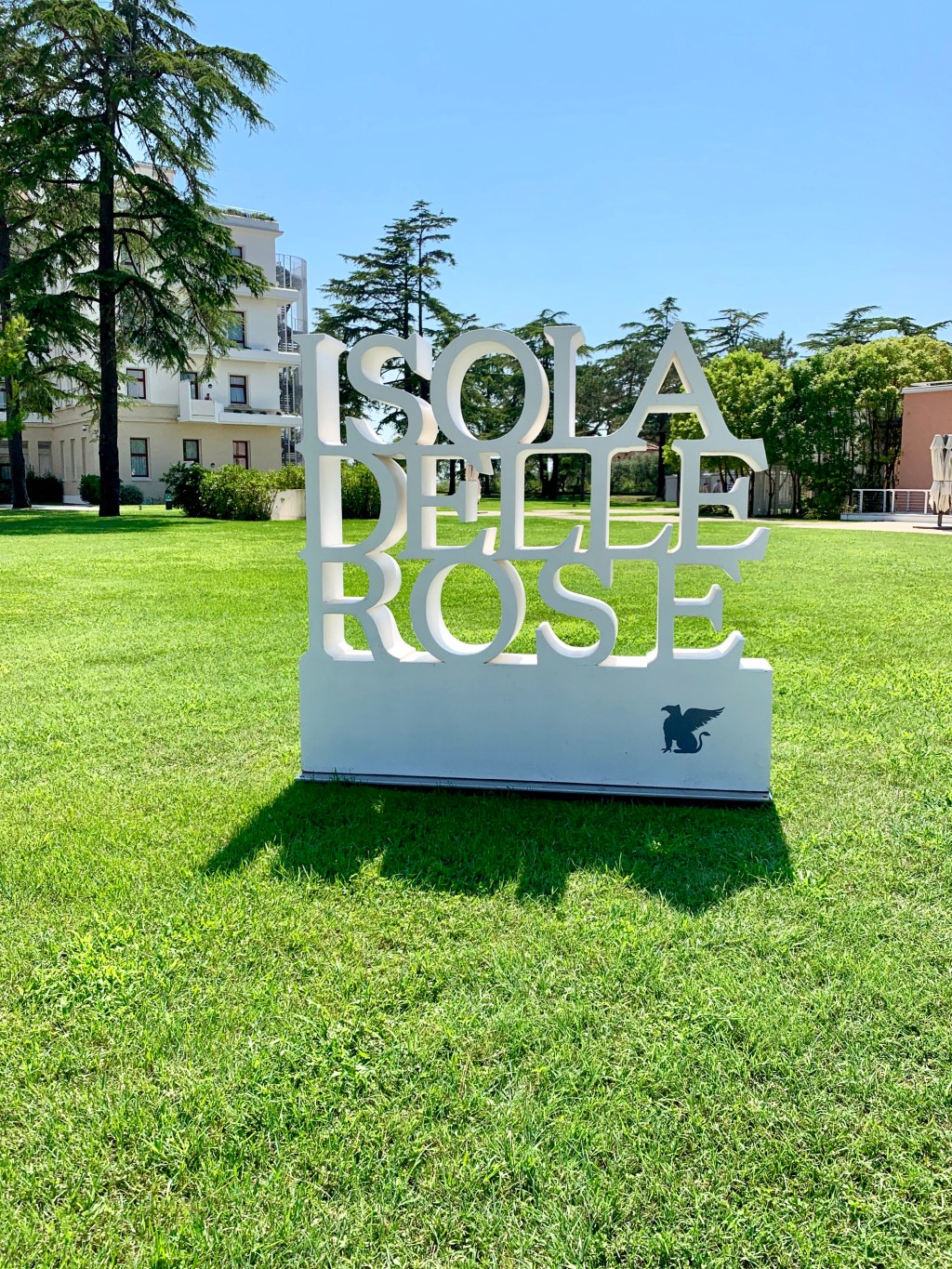 A large white sculptural sign reading ISOLA DELLE ROSE standing on a mown lawn between cedar trees, with the hotel buildings behind.