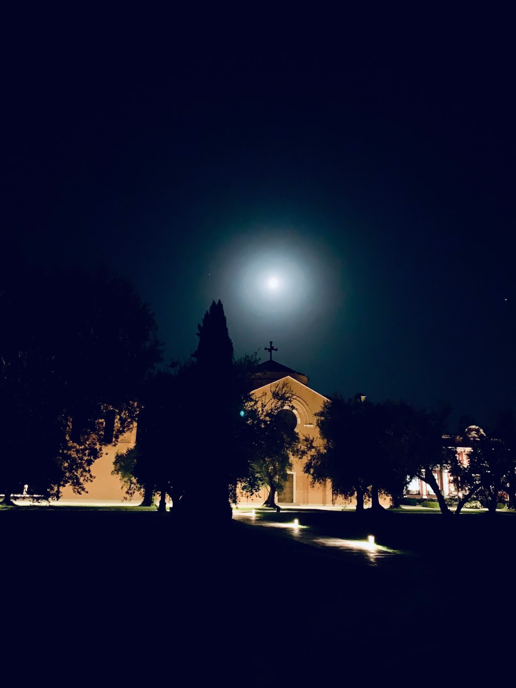 A full moon over the silhouette of a church and cypress trees at night, the grounds lit from below.