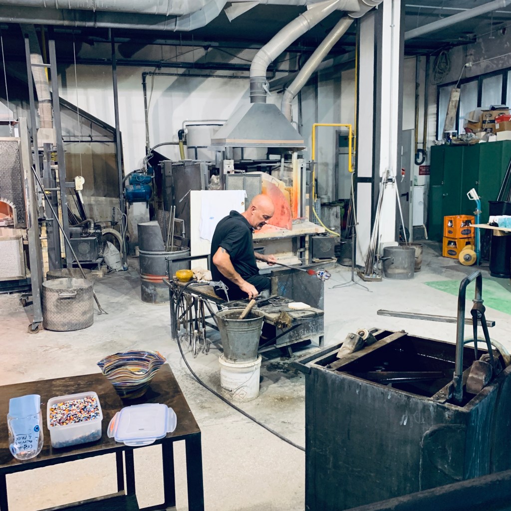 A glassblower at work in a Murano furnace workshop, seated at the bench with ducting and kilns around him and finished bowls on a trolley in the foreground.