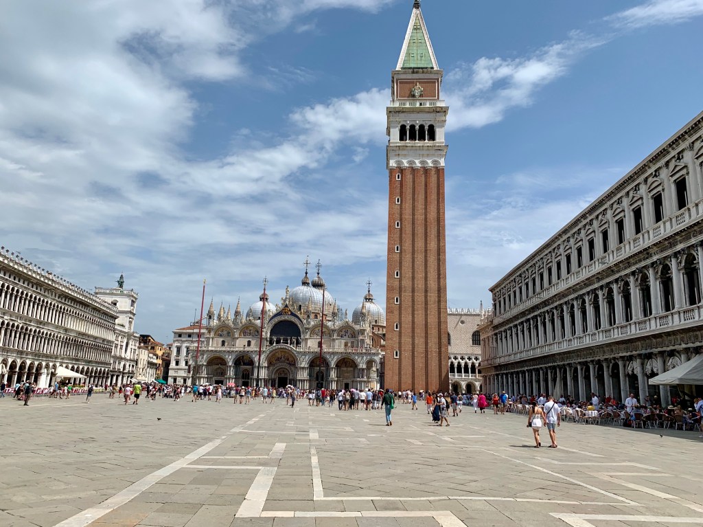 Piazza San Marco under a broken sky, the campanile rising at the centre with the basilica domes to its left and the arcades of the Procuratie running down both sides.