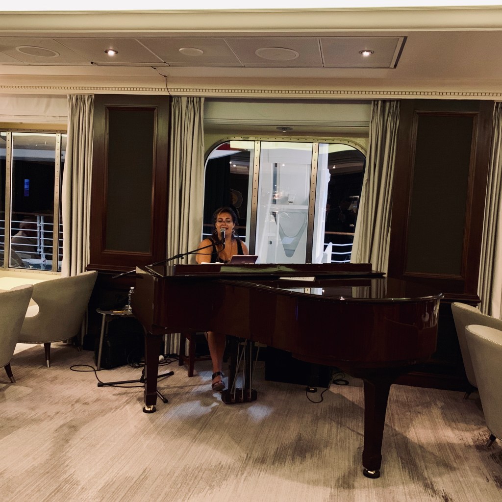 A woman singing at a grand piano in the ship’s lounge, surrounded by pale armchairs, with the sea visible through the windows behind.