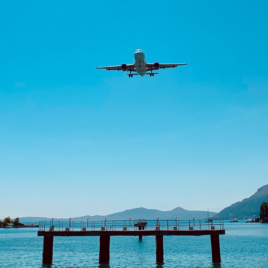 An aircraft on final approach into Corfu, low over the lagoon, with the wooded islet and the long jetty at Kanoni below it.
