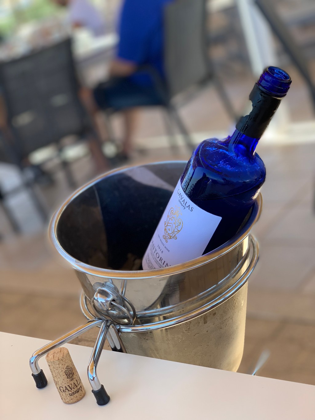 A cobalt blue wine bottle standing in a stainless ice bucket on a table, the cork resting on the cloth beside it.