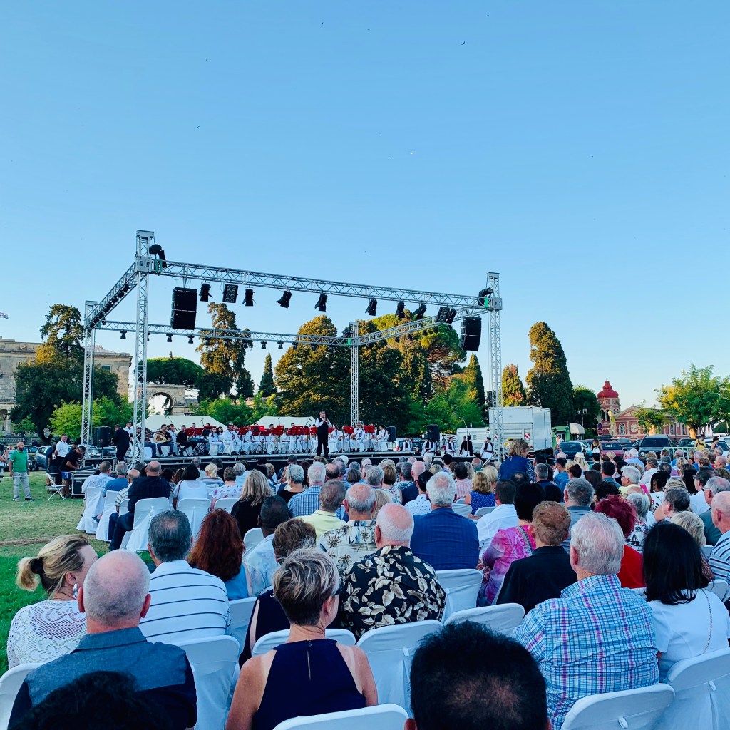 An open-air evening concert: rows of white chairs filled with an audience facing a lit stage where performers in white are lined up under a lighting rig.