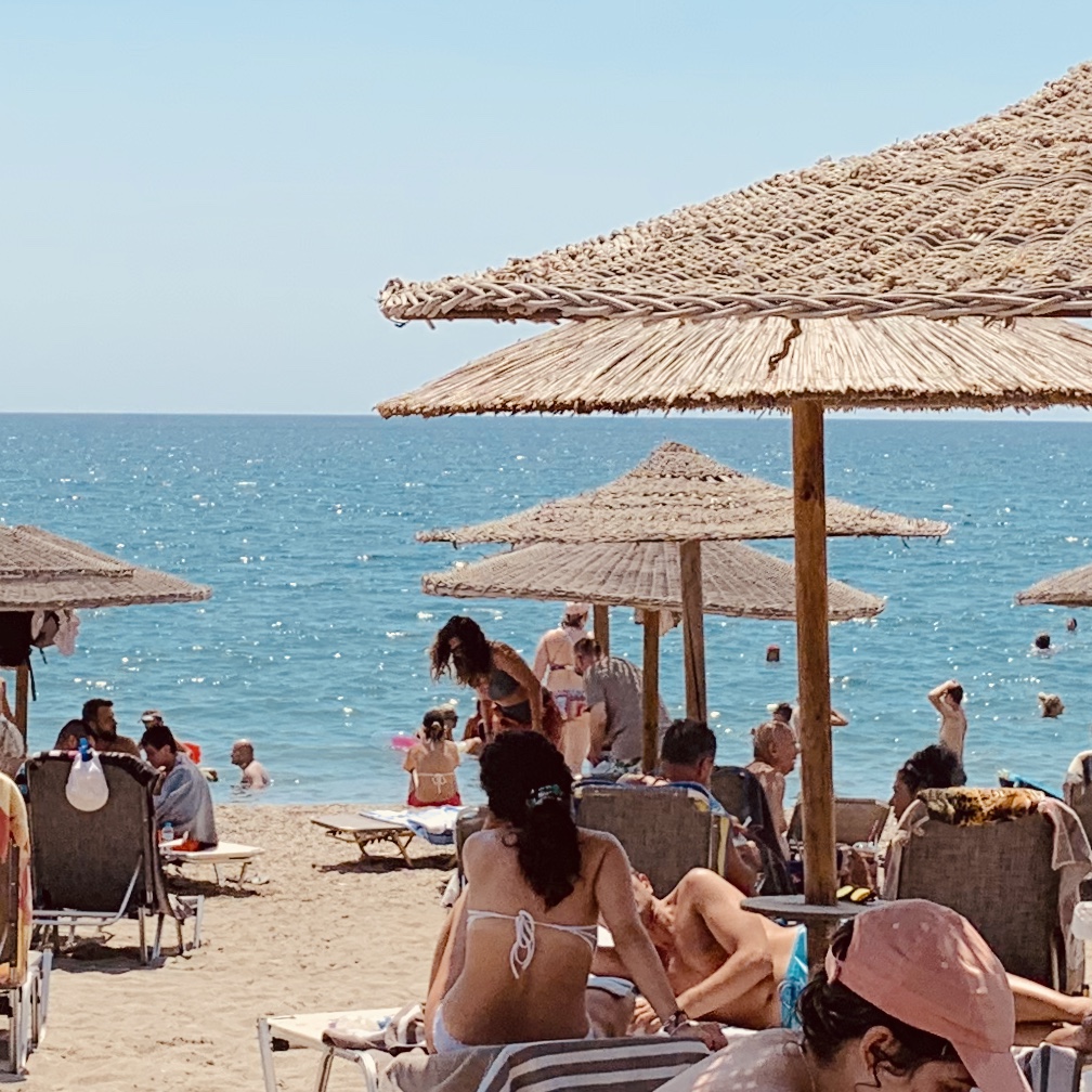 A Greek beach lined with rows of thatched straw parasols and sun loungers, bathers on the sand and flat blue sea beyond.