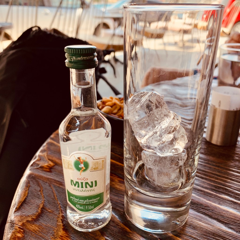 A small bottle of Mini ouzo beside a tall glass packed with ice on a varnished wooden table.