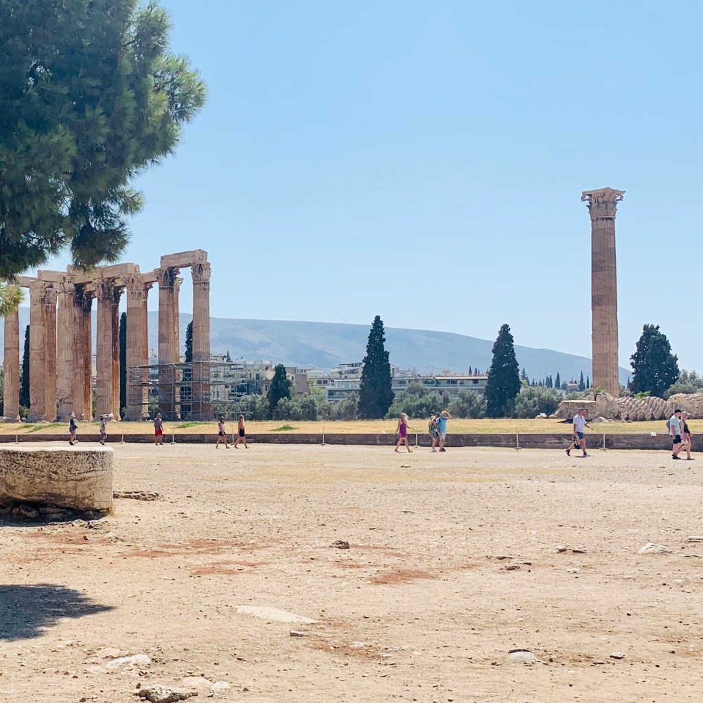 The surviving columns of the Temple of Olympian Zeus standing across a wide dusty archaeological site, a pine tree in the foreground.
