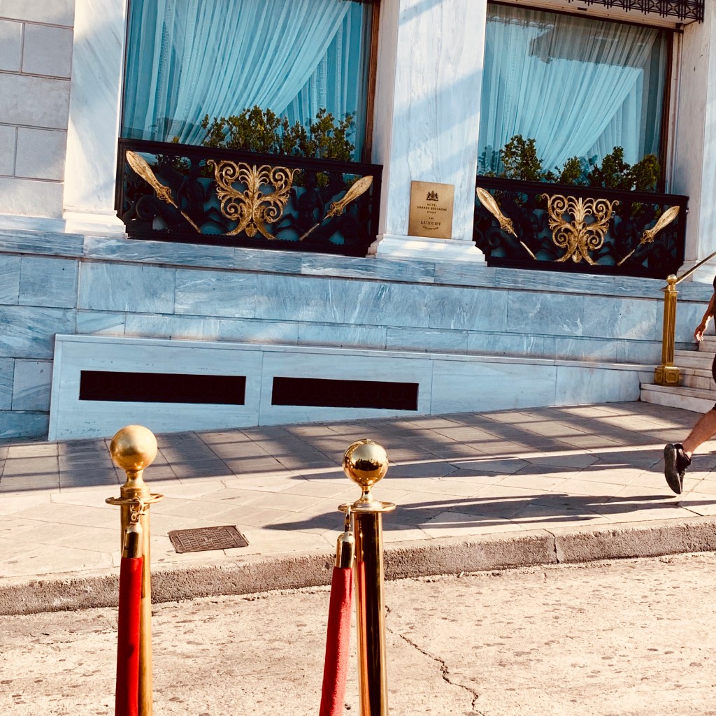 The marble facade of the Hotel Grande Bretagne with its blue and gold window boxes, red velvet ropes and brass stanchions along the pavement.