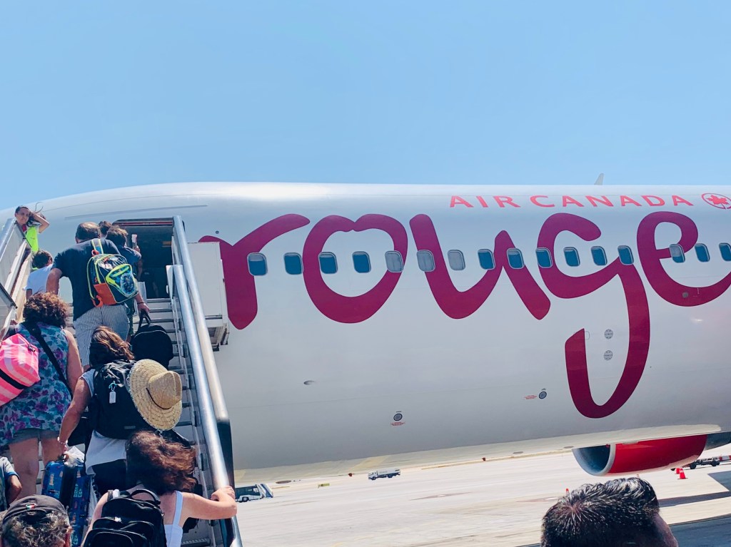 Passengers climbing the airstairs onto an Air Canada Rouge jet on the apron, the rouge lettering large along the fuselage.