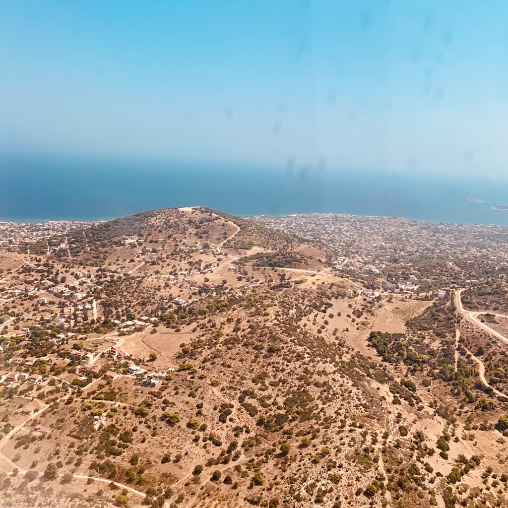 The Attic coastline from the air shortly after take-off: olive groves and scrub, scattered houses, and the sea running away to the horizon.