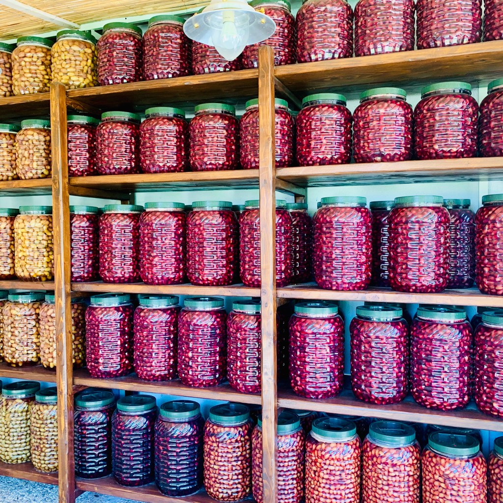 Wooden shelves stacked with rows of glass jars of preserved beans and pulses, backlit in a farm shop.