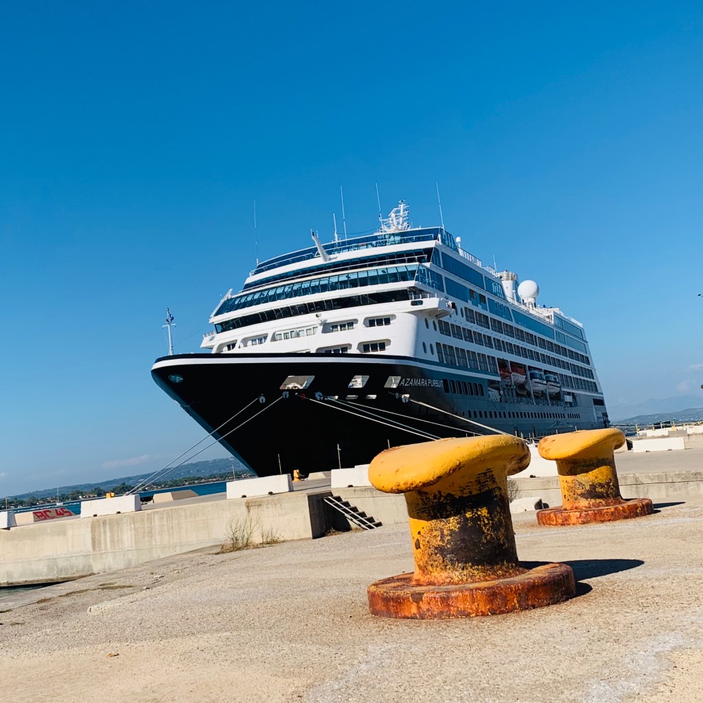 The Azamara Pursuit lying alongside the quay at Katakolon, seen past two rusted mooring bollards on the concrete pier.